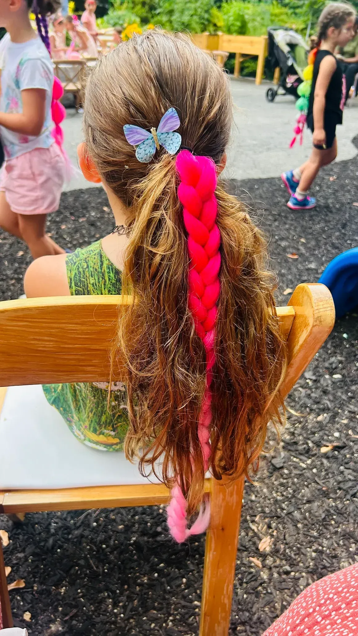 Child with brown, curly hair; pink braided extension, butterfly clip. Outdoors, on a chair.