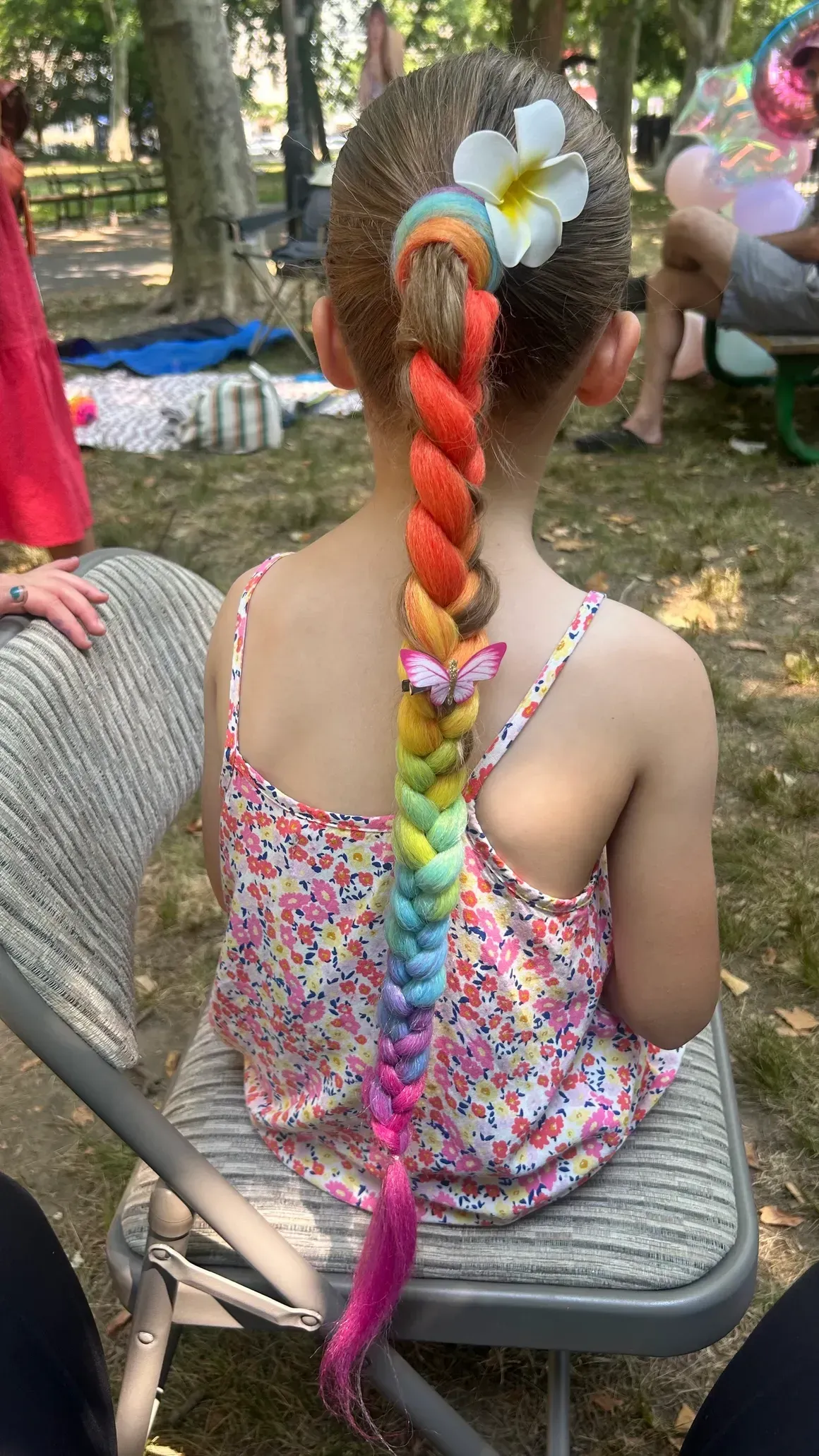 Young child with rainbow braid, white flower, and floral top, sitting outside.