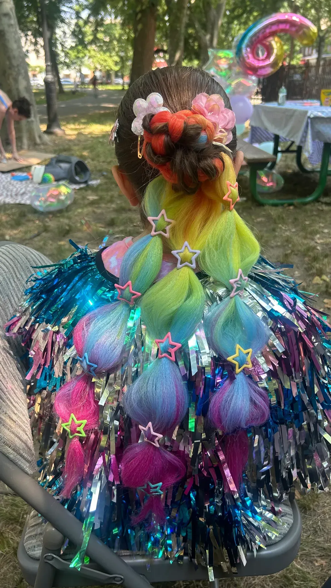 A person's hair styled with colorful decorations, including pom-poms, fringe, and flowers, outdoors.