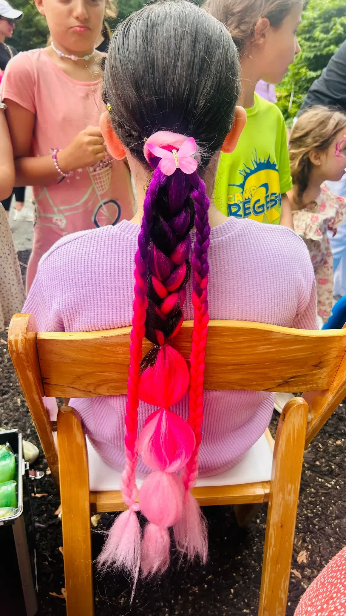 Girl's braided hair with ombre pink/purple extensions, seated on a wooden chair, at an outdoor event.