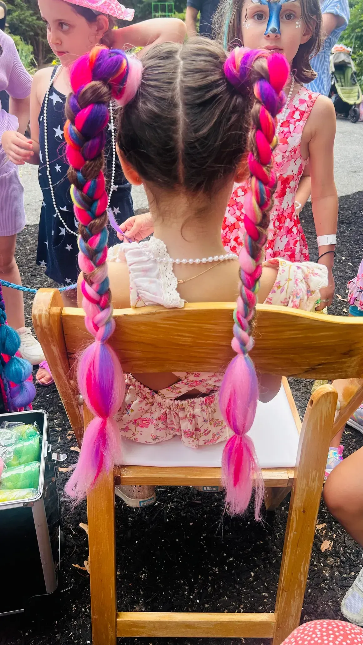 Girl with pink, purple, and blue braided pigtails seated in a wooden chair outdoors.