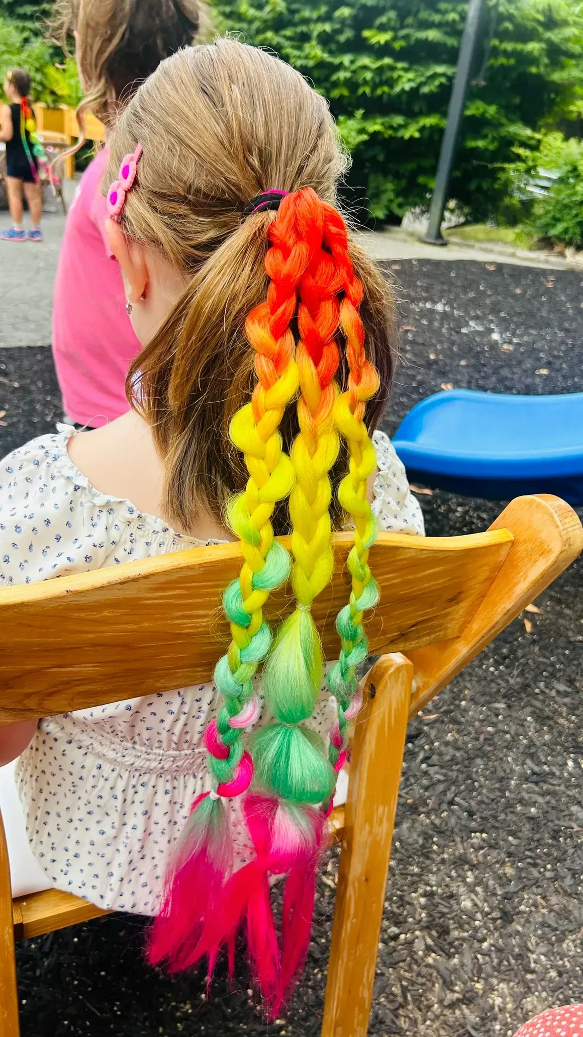 Girl with rainbow-colored hair extension braid. Sitting on a wooden chair. Outdoor setting.
