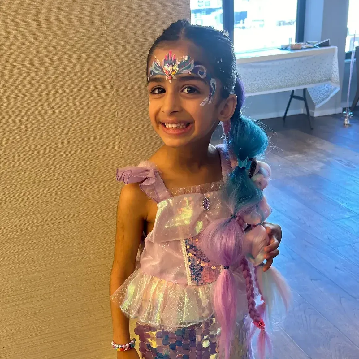 Girl in mermaid costume smiles, face painted. Holding colorful braided hair.