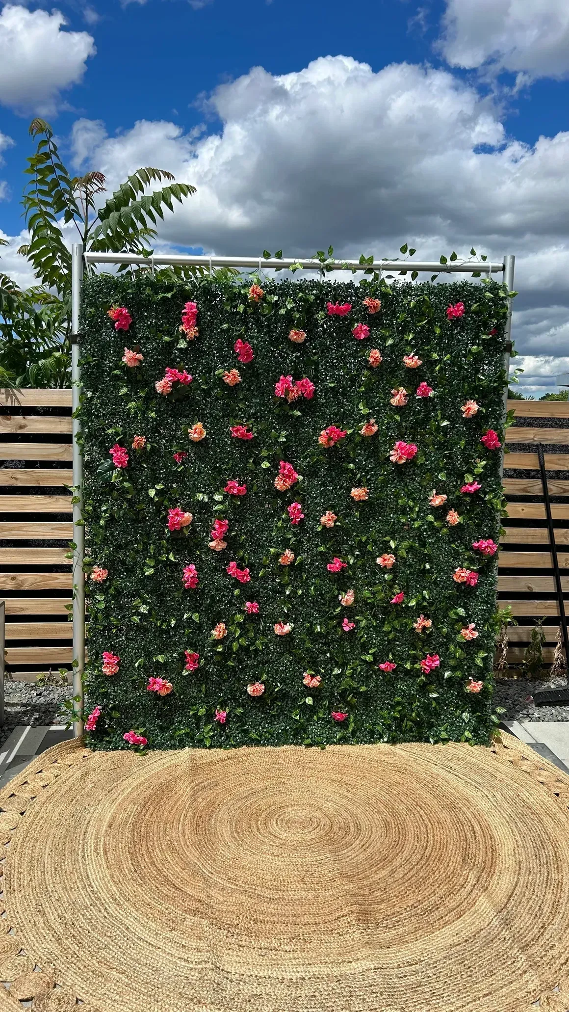 Square floral backdrop with pink flowers and lush greenery. Set on a straw mat against a blue sky.