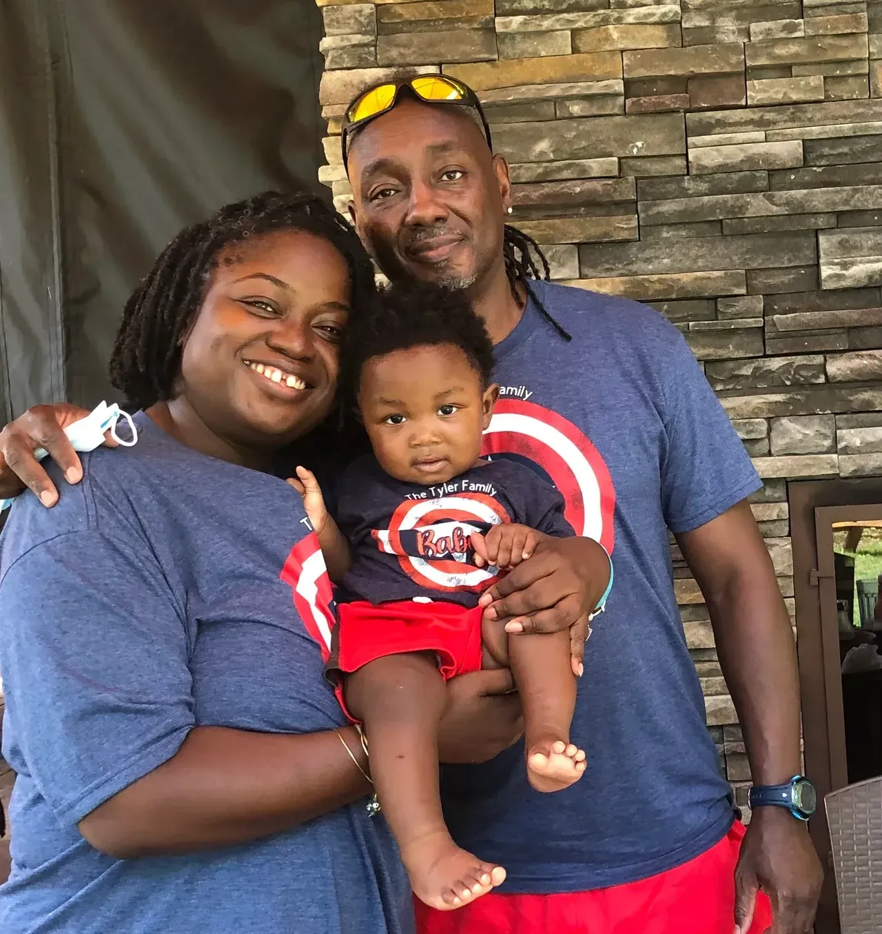Family of three in matching blue shirts and red shorts, smiling, posing in front of a stone wall.