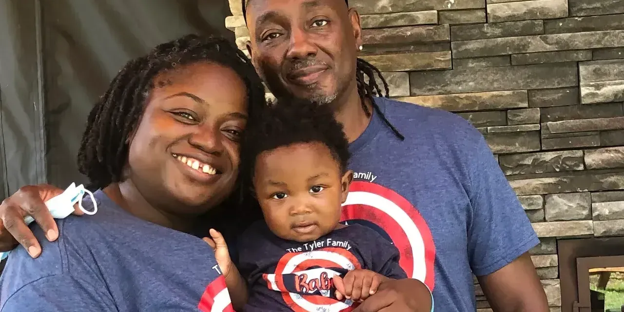 Family of three smiles together outdoors. A stone wall is behind them.