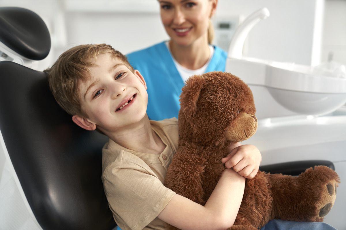 A young boy is sitting in a dental chair holding a teddy bear.