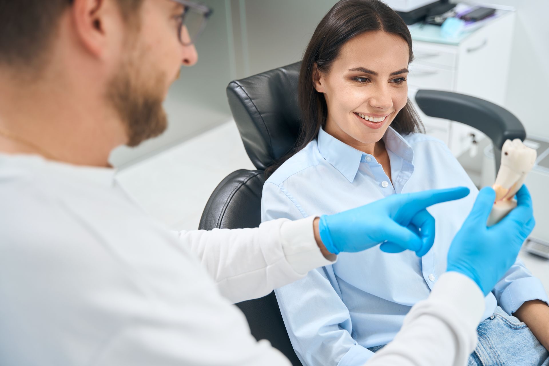 A woman is sitting in a dental chair while a dentist holds a model of a tooth.