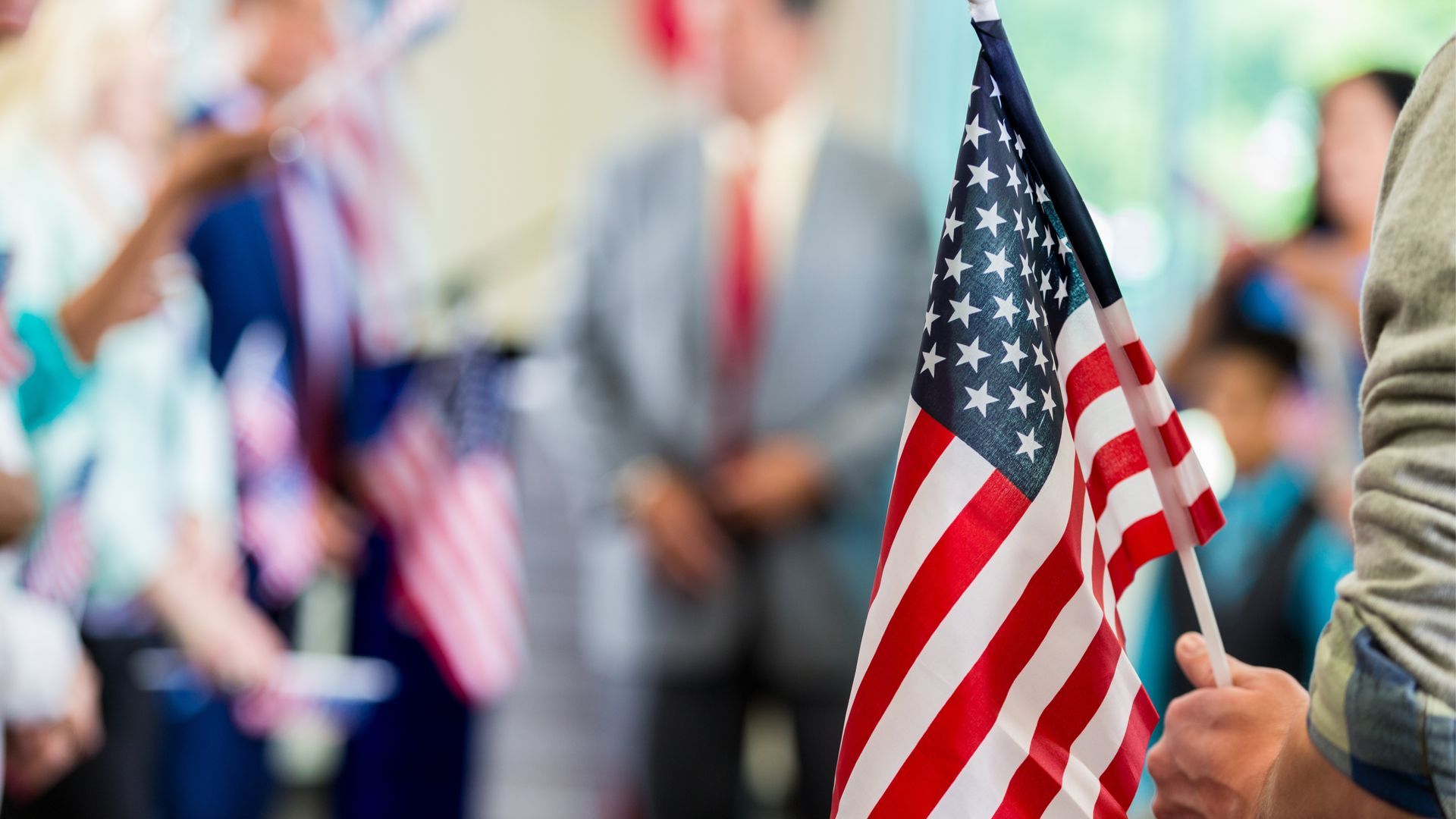 Person holding a small American flag; people in the background, likely at a public event.