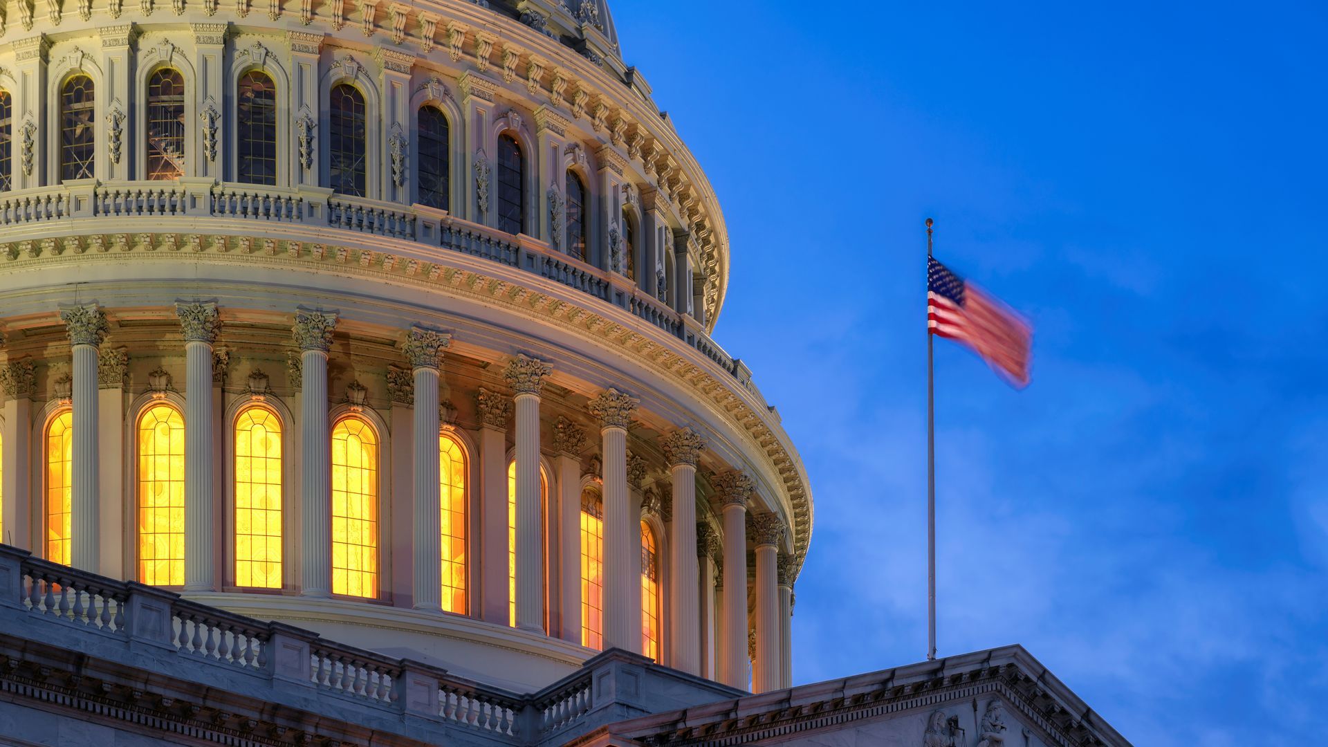 U.S. Capitol dome lit up at dusk, American flag waves in the wind.