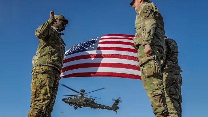 A group of soldiers standing next to an american flag and a helicopter flying in the background.