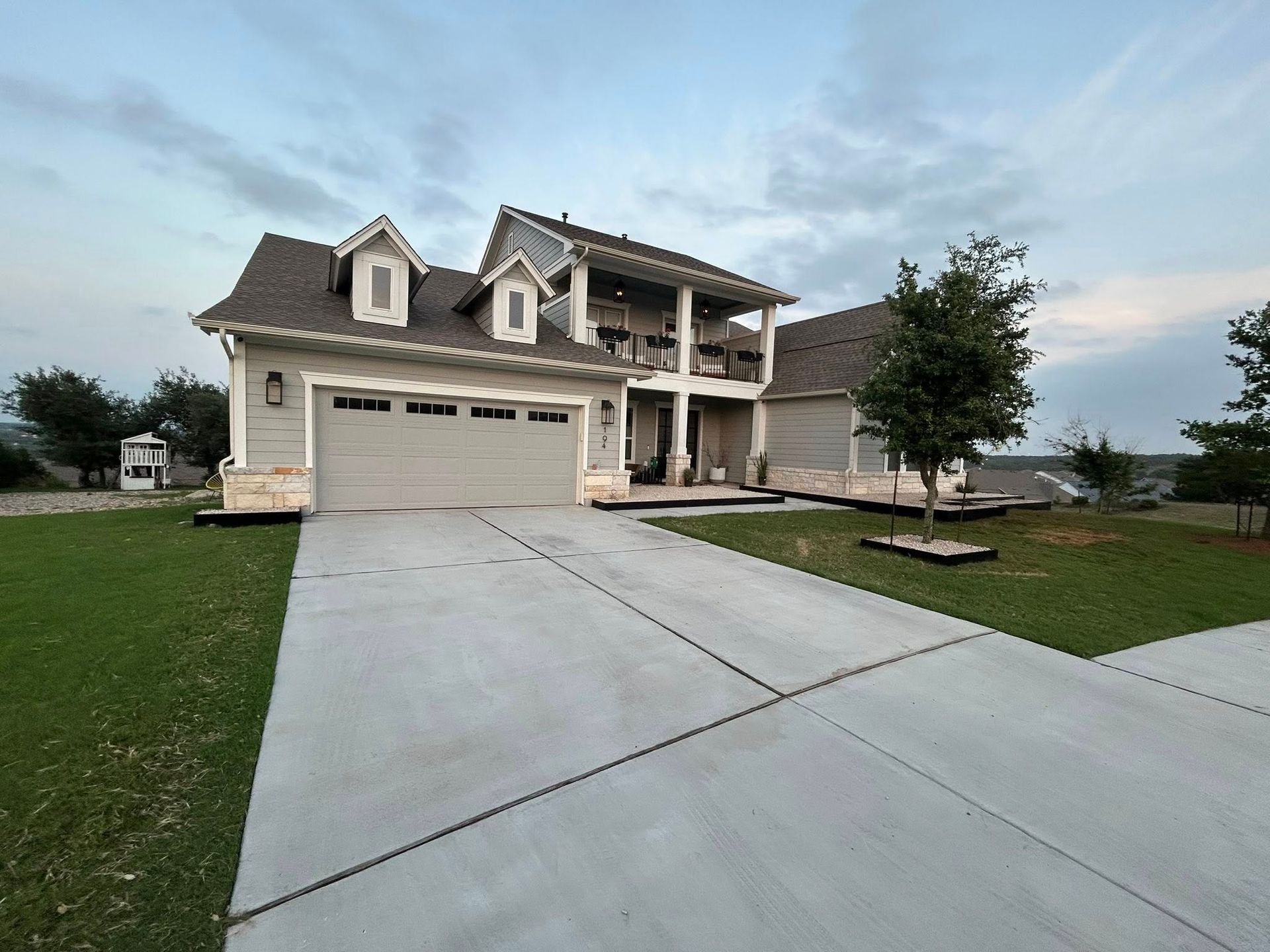 Two-story house with a gray exterior, attached garage, balcony, and large concrete driveway under a cloudy sky.