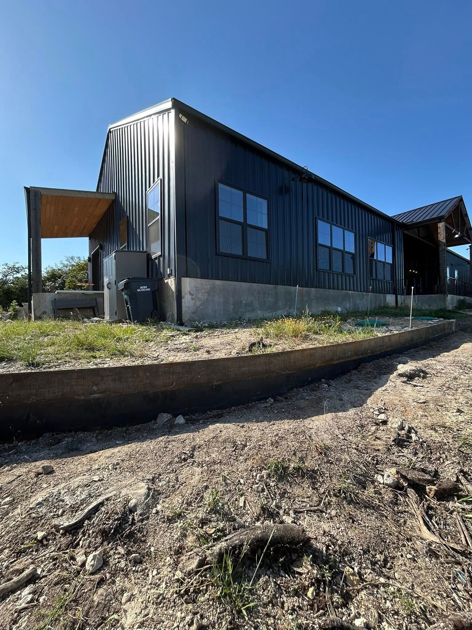 Black metal building with multiple windows, a porch, and a trench in front, set against a blue sky.