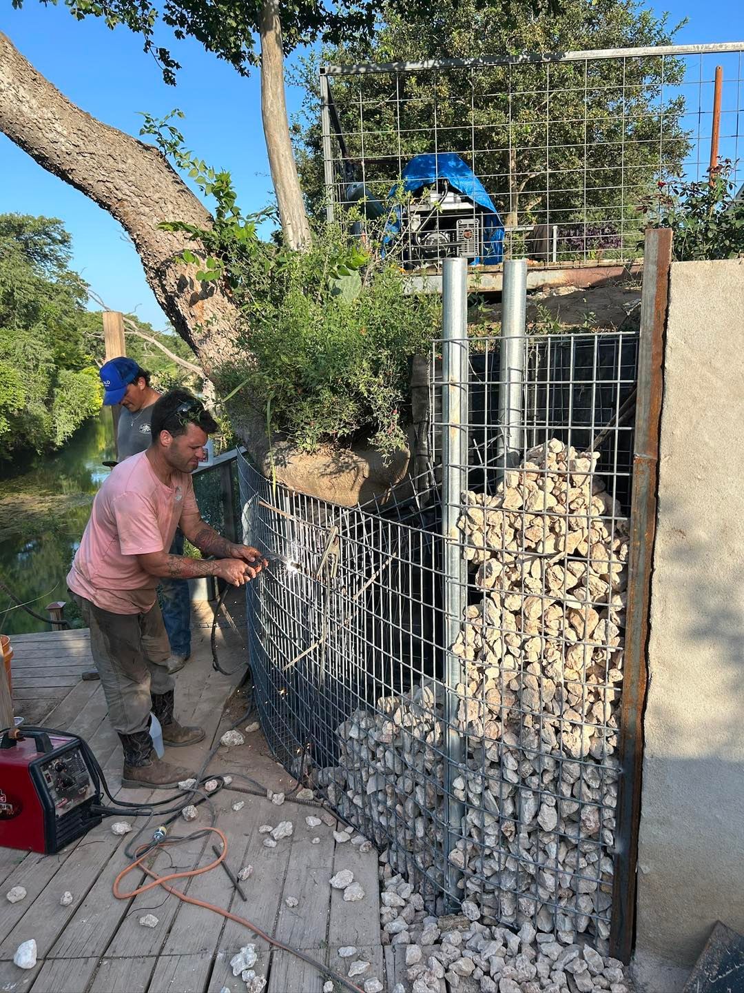 Two men constructing a gabion wall; one welding, the other securing wire. Rocks fill wire mesh cages next to a deck, near a tree.