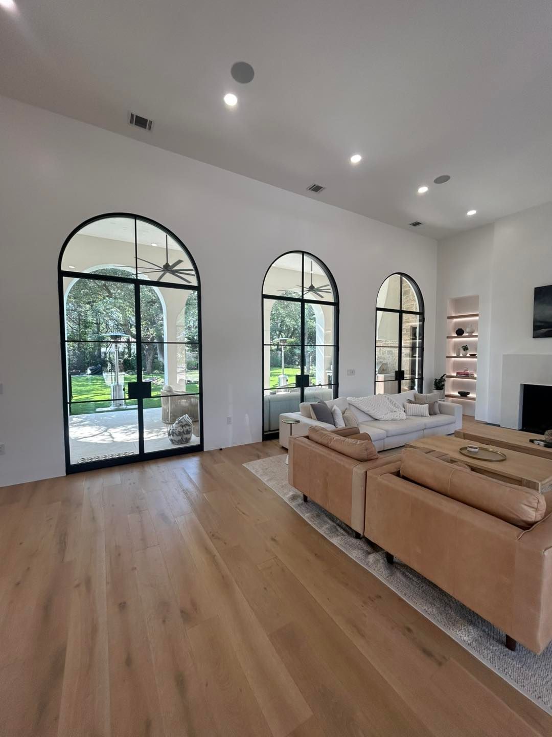Living room with arched black framed windows and wood floors; beige leather couches and white walls.