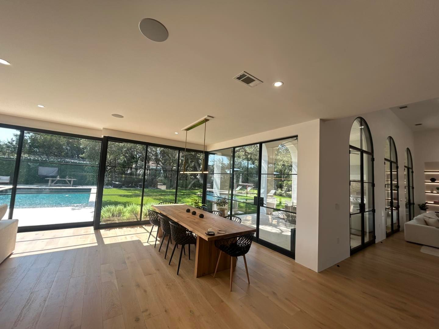Spacious dining room with a long wooden table, black chairs, and floor-to-ceiling windows overlooking a pool and yard.