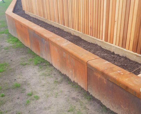 Rusty metal planter box along a wooden fence, filled with dark soil, on grassy ground.