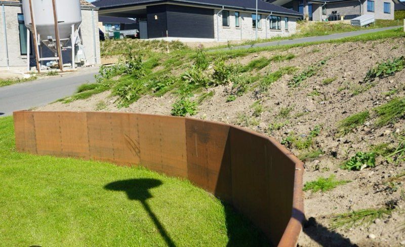 Rusty metal retaining wall in a yard, separating grass from a hillside with sparse vegetation and buildings in the background.