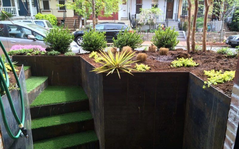 Steps with green turf leading to a garden bed. Metal retaining walls hold soil with plants; cars and houses in background.