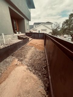 Construction site with a long, brown metal retaining wall beside a path. House in the background.