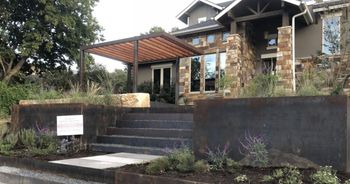 Stone house with steps leading to the entrance, brown pergola over the door, and landscaped yard.