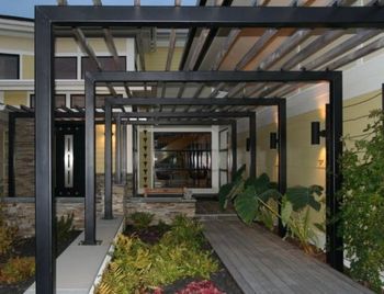 Black metal pergola frames walkway to a modern home entrance with plants.