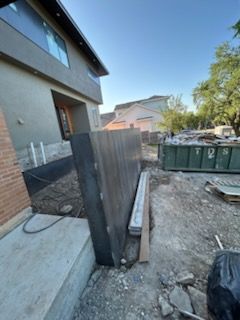 Construction site: dark wood fence, building with stucco, green dumpster, and dirt.