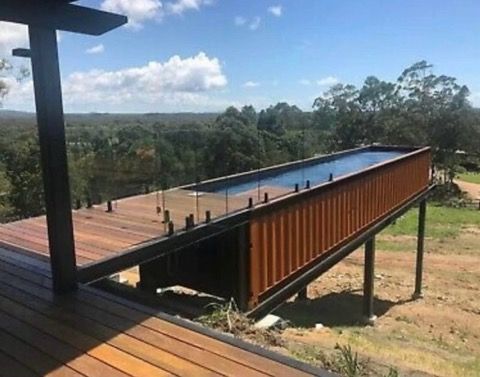 Elevated shipping container pool with wood deck and glass railing, overlooking a green landscape.