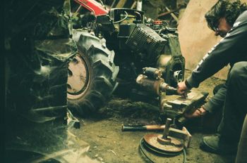 Person repairs machinery in a dim, cluttered space. A tractor wheel is visible on the left.