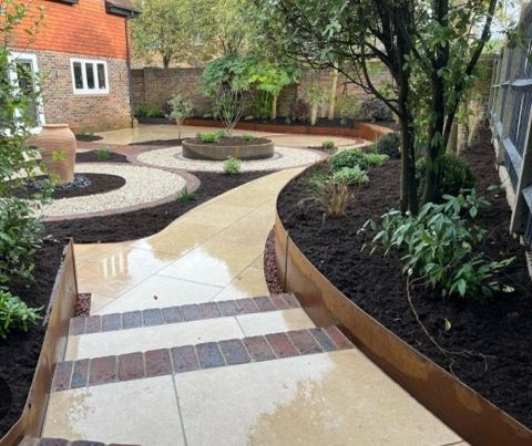 A garden with stone pathways, circular gravel beds, and a brick house in the background.
