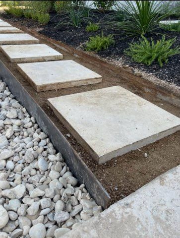 Concrete stepping stones in a dirt path with a metal border, bordered by white rocks and plants.