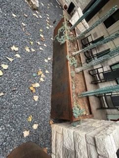 Long, copper planter with trailing greenery next to exterior stone steps, gravel ground, and patio seating.