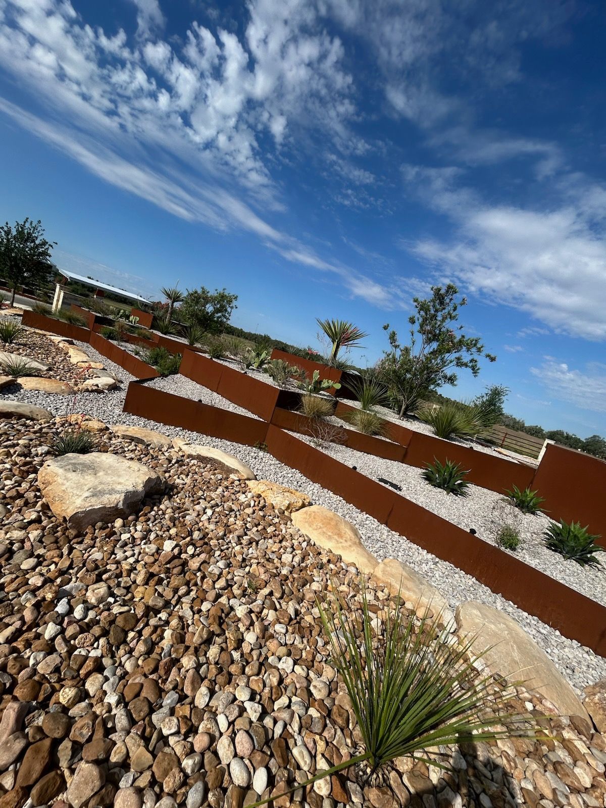 Terraced garden with rusty metal borders, gravel, rocks, and plants under a blue sky.
