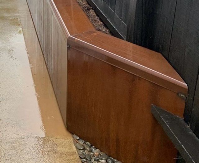 Brown metal planter box next to a fence, filled with rocks and dirt.