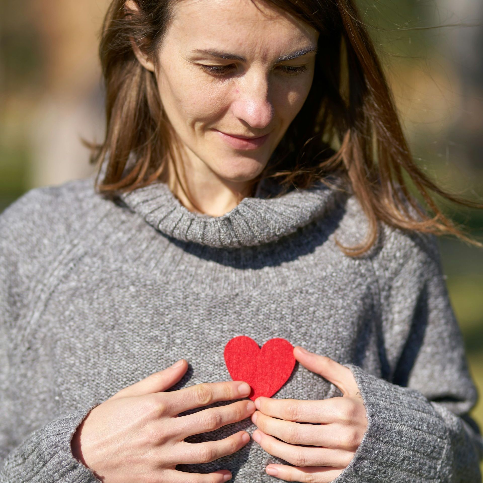 A woman in a grey sweater is holding a red heart in her hands.