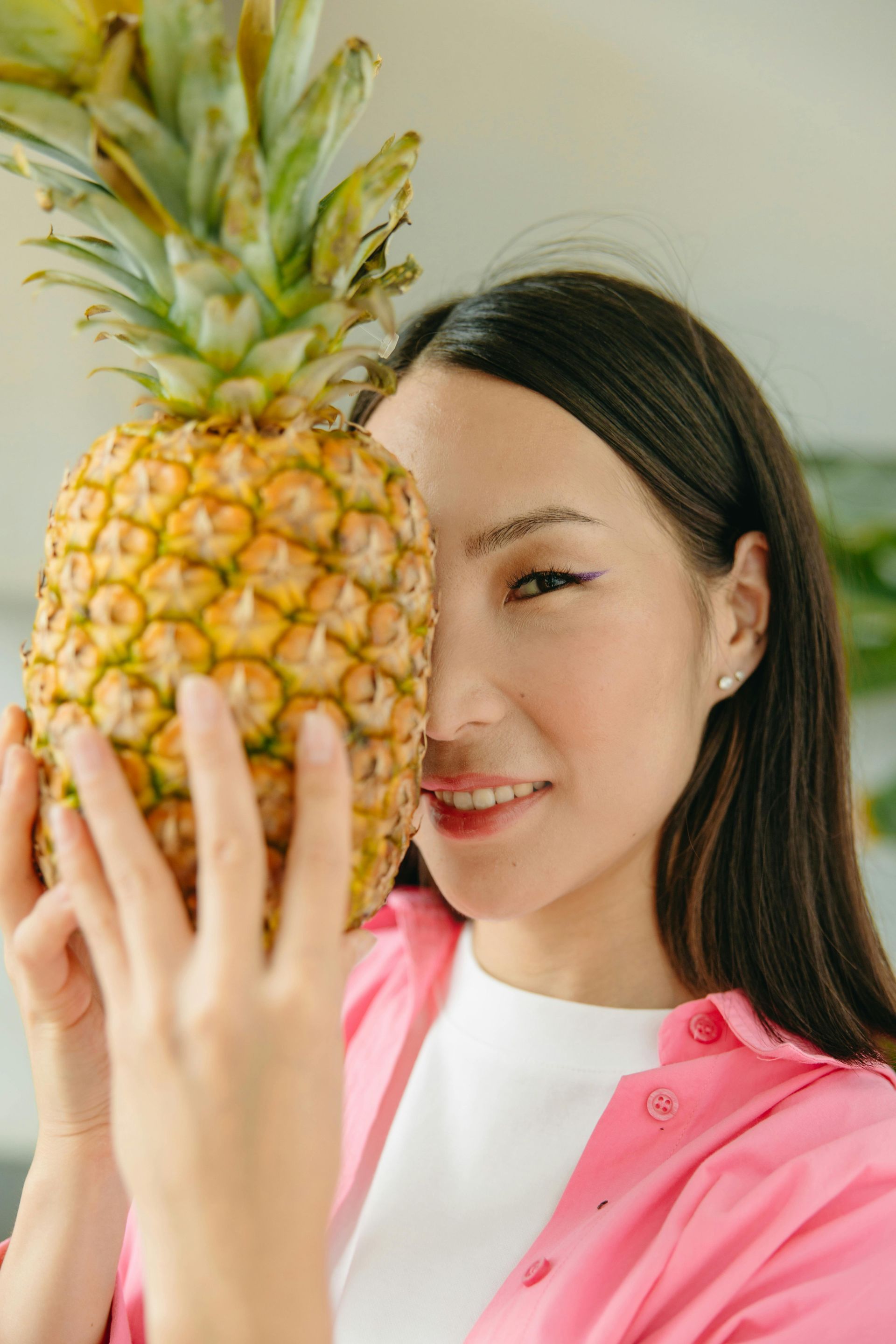 A woman in a pink shirt is holding a pineapple in front of her face.
