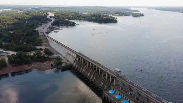 Aerial view of a dam and reservoir with trees and a road visible in the background. Blue water, concrete, and nature.
