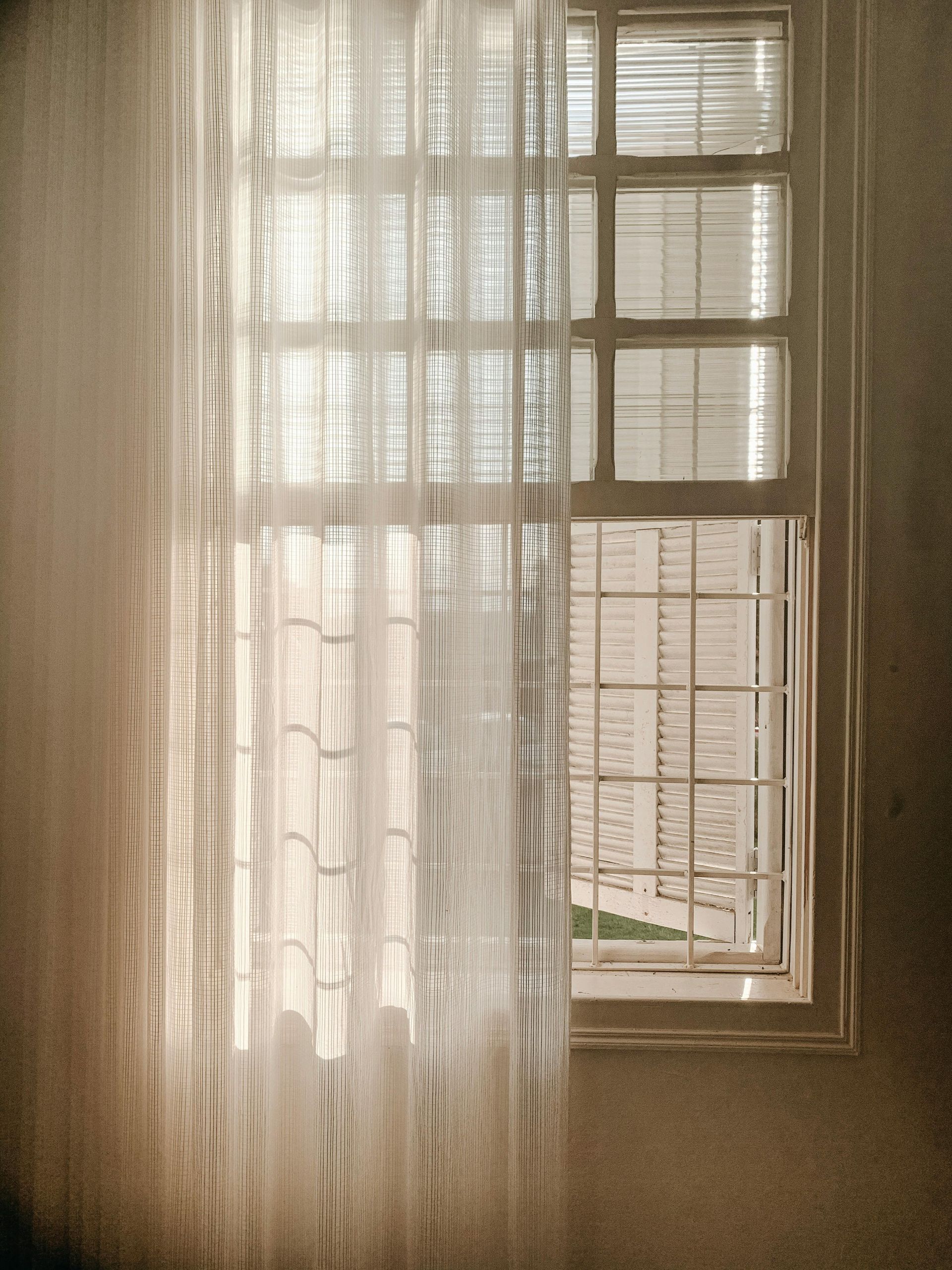 Window with sheer curtain, sunlight filtering through, illuminating a building in the background.