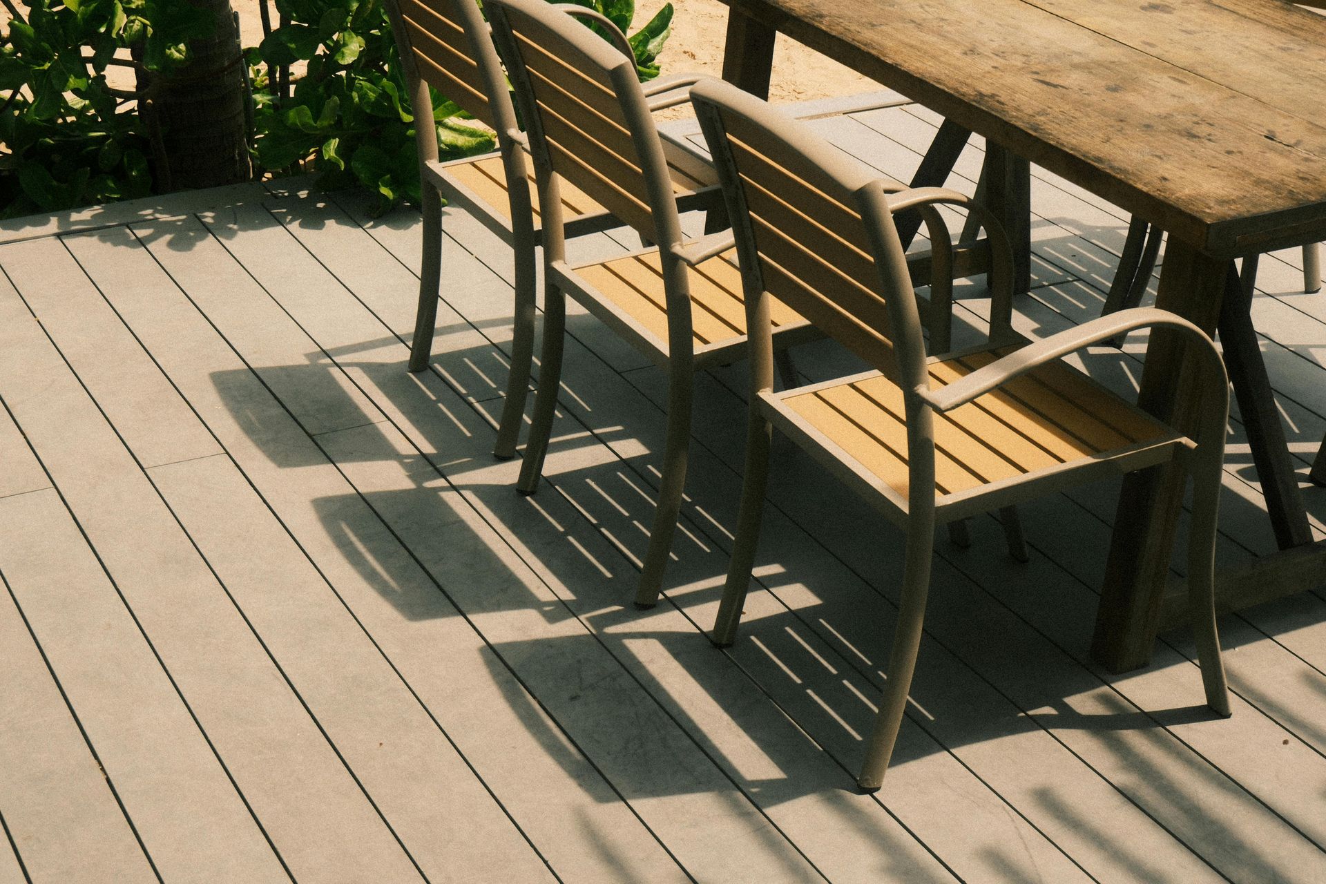 A wooden dining table and three matching chairs sit on a light-colored outdoor deck next to some green plants.