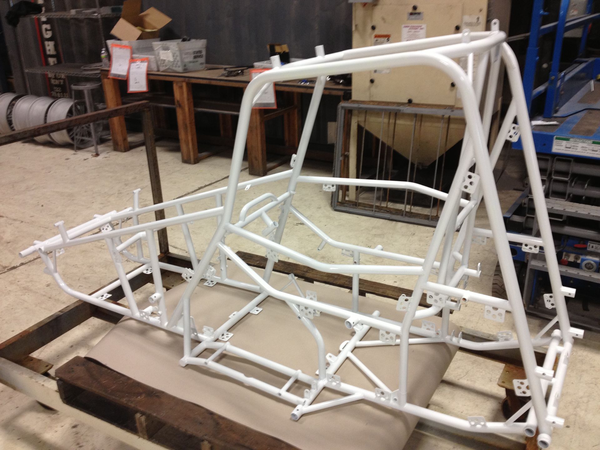 White-painted, tubular steel frame, likely a vehicle chassis, resting on a wooden pallet in a workshop.