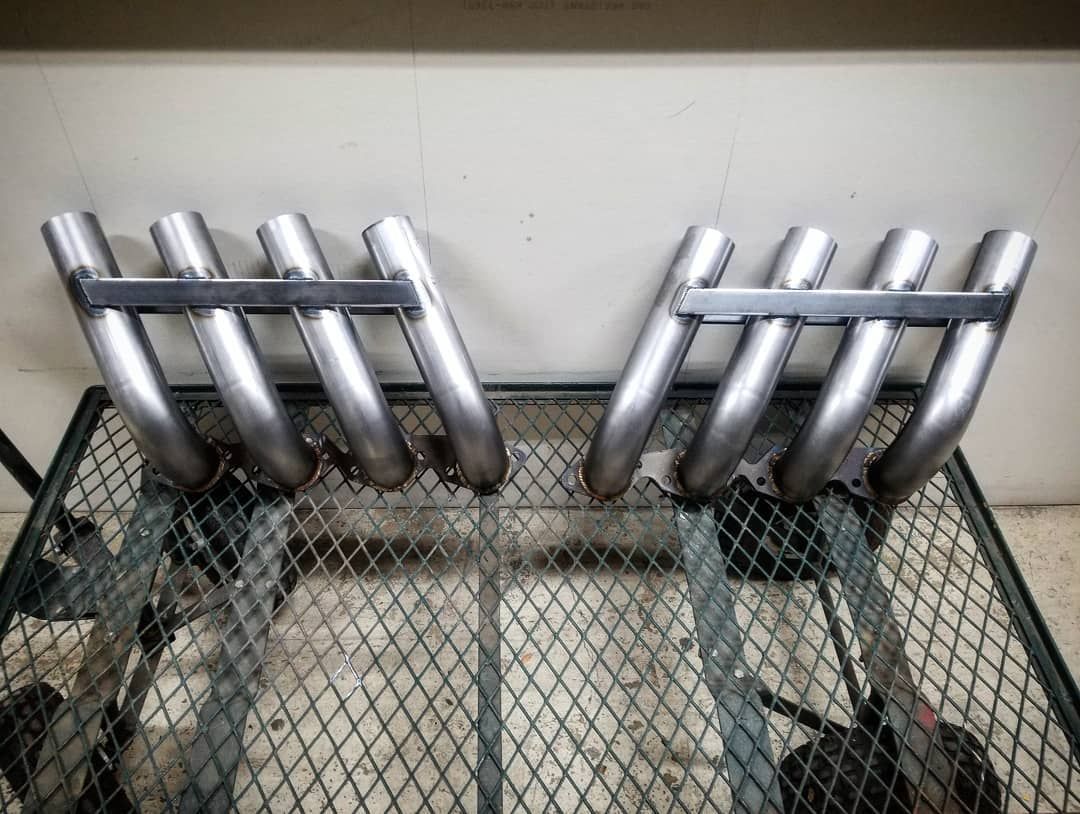Two silver exhaust headers, newly fabricated, resting on a metal grate.