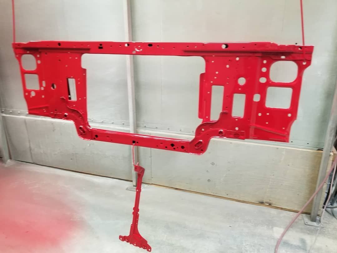 Red painted car part, likely a front support panel, hanging in a spray booth.