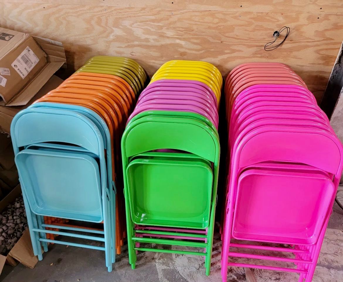 Stacks of colorful folding chairs: blue, orange, yellow, pink, and green against a wooden wall.