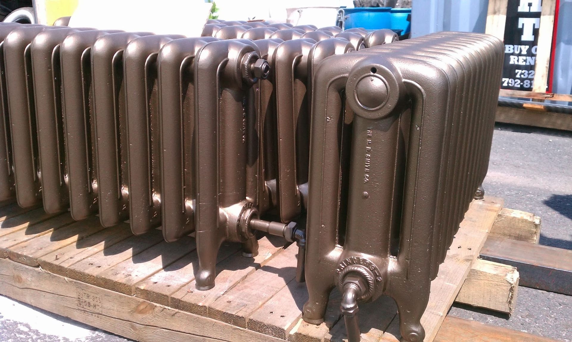 Several old cast iron radiators stacked on a wooden pallet, painted a bronze color, outdoors.