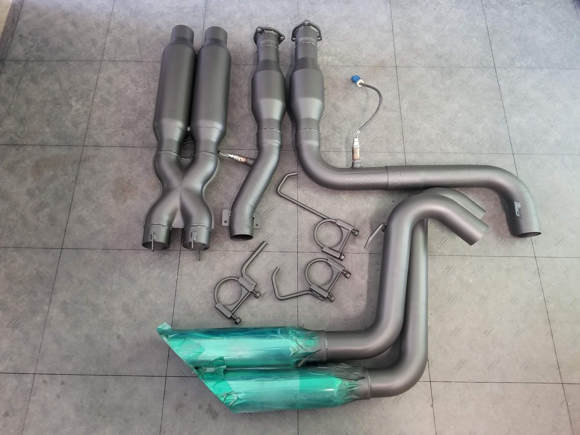 Gray car exhaust system components laid out on a tiled floor.