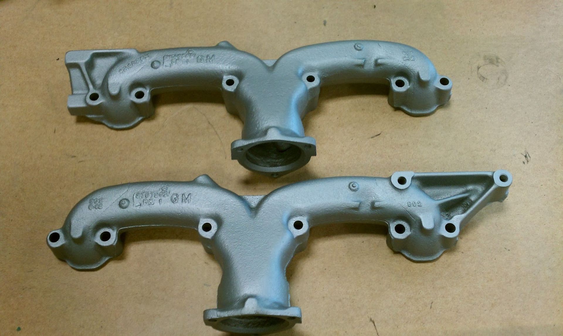Two gray cast-iron exhaust manifolds, side by side, on a wooden surface.
