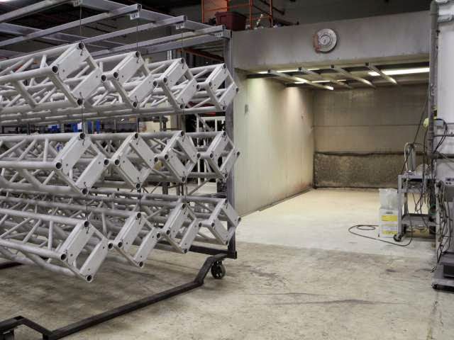 Metal trusses on a rack next to an industrial paint booth with open doorway.