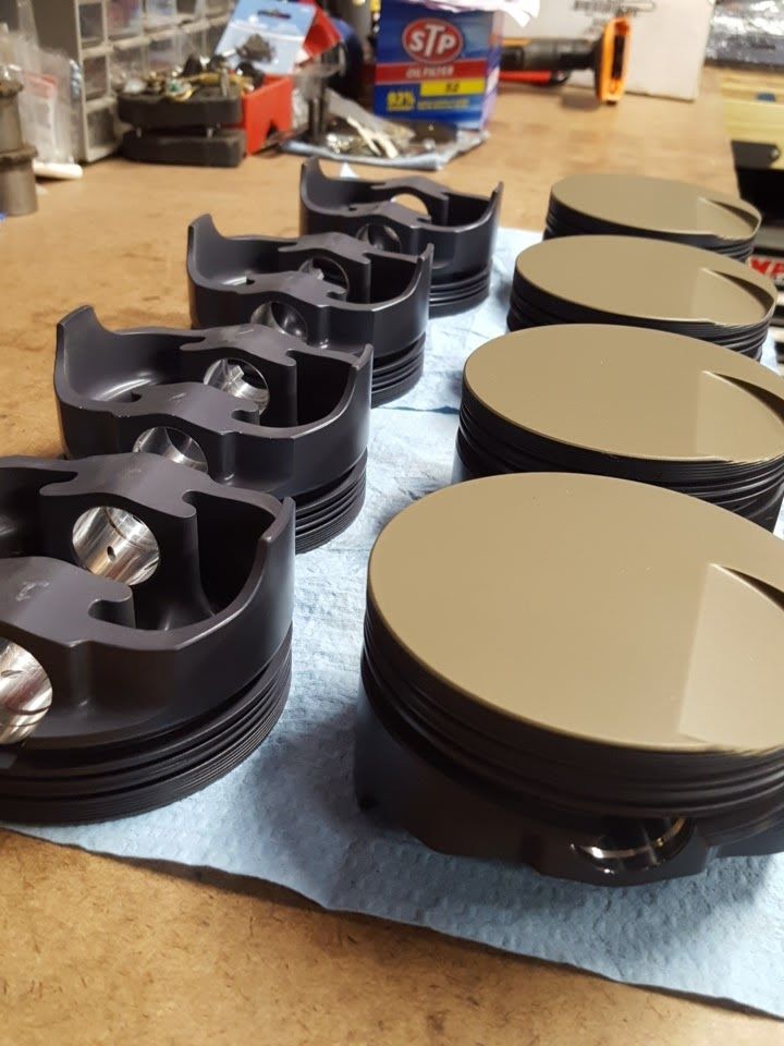 Eight new engine pistons lined up on a blue paper towel. The pistons are black and tan.