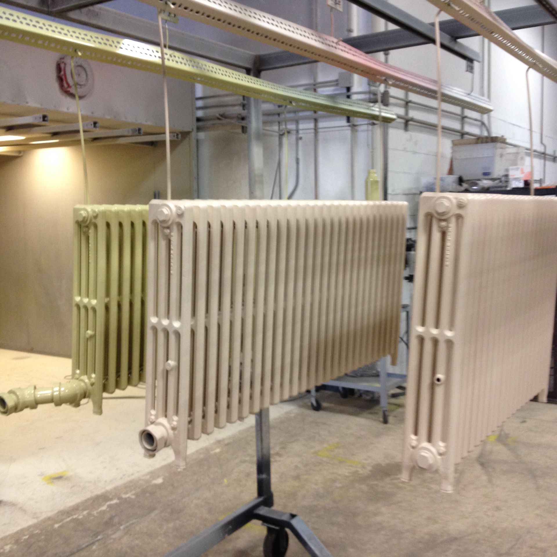 Three painted radiators hanging from a factory overhead track. Two are beige, one is green.