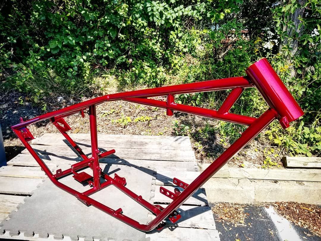 Red motorcycle frame outdoors on gray flooring, in front of leafy green foliage.
